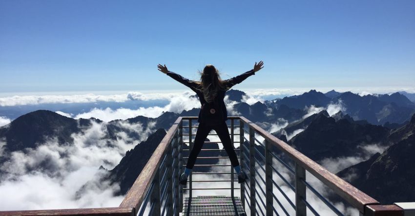 Adventures - Person Standing on Hand Rails With Arms Wide Open Facing the Mountains and Clouds